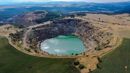 Aerial view of Tsar Asen Mine, Bulgaria – a former open-pit mine now filled with striking turquoise water. The steep terraced slopes and surrounding fields reflect the site's industrial past and natur