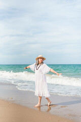 Carefree senior woman in white dress and straw hat enjoying freedom walking barefoot on the beach