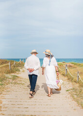 Elderly couple walking on a beach path enjoying retirement with a picnic basket and summer outfit