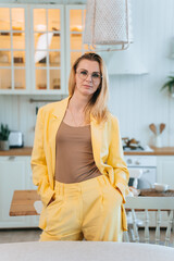 A confident woman in a bright yellow suit stands in a cozy, modern kitchen, exuding elegance and poise.