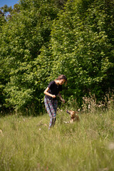 Fototapeta premium Happy young woman playing with dog in dandelion field and trees in the background 