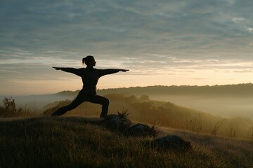 Woman practices warrior II pose on a hilltop during sunrise with misty landscape