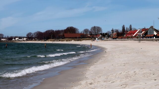 Empty Baltic Sea beach with gentle waves and a coastal village in the background. Early spring atmosphere with calm water and quiet seaside town life.