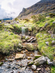 A small waterfall cascades over rocks and vegetation in a picturesque landscape. This is along the popular hiking trail of Carrauntoohill in County Kerry, Ireland, with a mountain in the background.