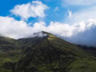 View of rugged Carrauntoohill mountain partially shrouded in clouds against a bright blue sky in County Kerry, Ireland.