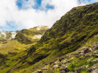 Hills covered in green vegetation and scattered rocks form an Irish mountain landscape. Mist and clouds roll through the valleys near Carrauntoohil along the hiking trail in County Kerry.