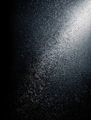 Black matte textured background with floating dust particles illuminated by soft light in the top right corner, creating a deep contrast between black and white. This abstract design emphasizes textu