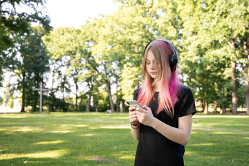 Young girl with pink hair wearing headphones is focused on using smartphone in a lush green park,...