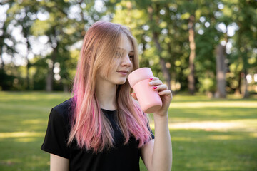 Fototapeta premium Young woman with pink hair is enjoying a drink from a pink cup while standing in a lush green park, embodying a relaxed outdoor lifestyle