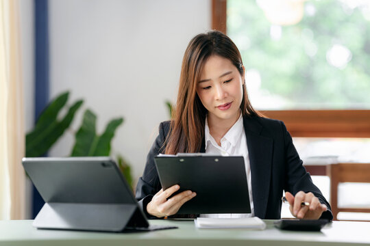 A woman in a business suit is sitting at a desk with two laptops and a clipboard