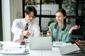 Asian man and European woman working on a laptop discussing business data analytics and financial charts in office for planning strategy together