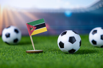 Miniature Mozambique flag with multiple soccer balls on green grass turf, symbolizing national team participation and global football competition within a stadium arena