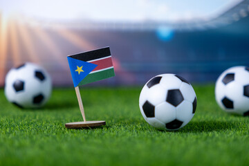 South Sudan flag miniature on artificial green turf with small soccer balls, representing national football team participation in stadium competition