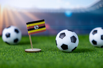 Small Ugandan flag proudly stands with miniature soccer balls on green artificial grass, symbolizing national pride for international football tournaments
