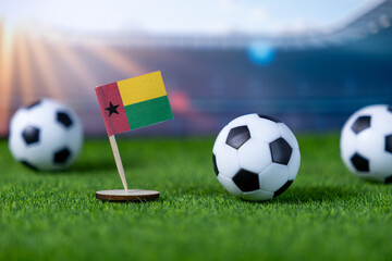 Vibrant Guinea-Bissau flag standing alongside miniature soccer balls on lush green grass emphasizes international football competition and national team spirit in a stadium
