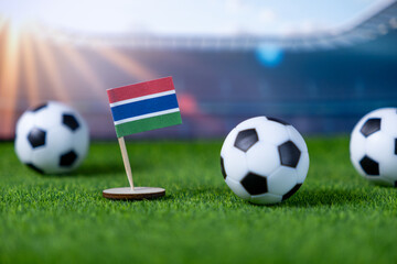 Miniature Gambia flag on green grass with soccer balls, blurred stadium lights, symbolizing national pride for international football competition event