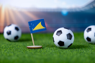 Small Tokelau flag stands firmly on green artificial grass with soccer balls, representing global...