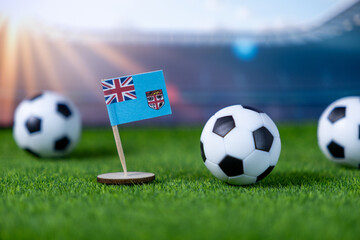 Small Fiji flag standing proudly on green football pitch alongside soccer balls, symbolizing national pride, sports spirit, and global competition