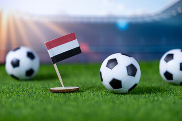 A miniature national flag of Yemen proudly stands on a lush green football pitch with soccer balls, symbolizing international sports competition and national team spirit