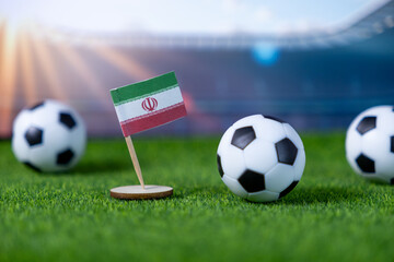 Miniature Iranian flag on a green football pitch with soccer balls, representing international sport, national pride, and competition in stadium setting