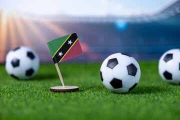Saint Kitts and Nevis national flag proudly stands with miniature soccer balls on vibrant artificial grass, set against a blurred stadium, symbolizing global sports event