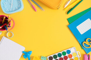 Colorful assortment of school stationery with empty space on bright yellow background including pencil case, alarm clock, notebooks, pencils, paints and more, top view