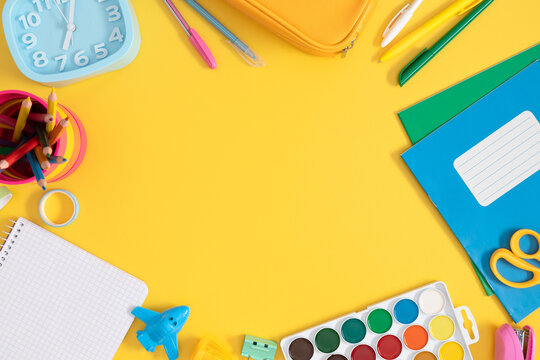 Colorful assortment of school stationery with empty space on bright yellow background including pencil case, alarm clock, notebooks, pencils, paints and more, top view
