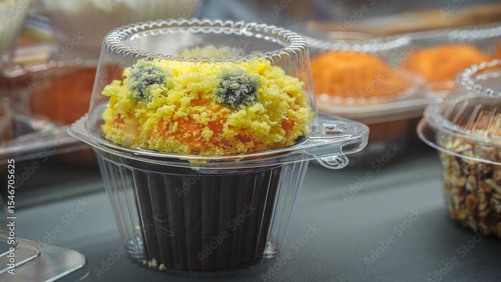 Poster cupcake in a plastic container with a yellow frosting and a blue decoration. the cupcake is sitting 