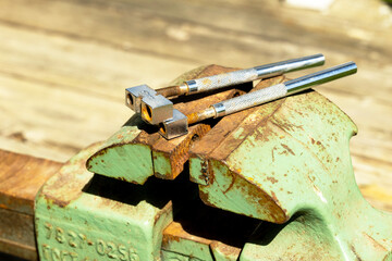 A rusty green vise holds two metal calipers on a wooden surface. The scene captures tools used for precision work in a workshop setting.