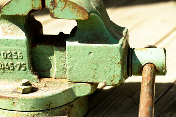 A close-up of a vintage green vise on a wooden surface. The vise shows signs of wear and rust, highlighting its industrial design and functionality.