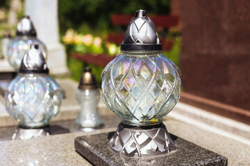Glass grave candle on tombstone. Shiny lantern with metallic base. Decorative cemetery lamp in sunlight. Memorial light at cemetery. Symbol of memory and respect. Death and remembrance concept.