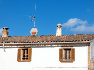 House, with traditional weathered tiled roof, older style, a warm climate. Chimney top on the roof. Satellite Dish a television antenna, windows with open wooden shutters, blue sky with white clouds. 
