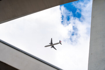 A commercial jetliner flies overhead, perfectly framed by modern urban architecture against a...