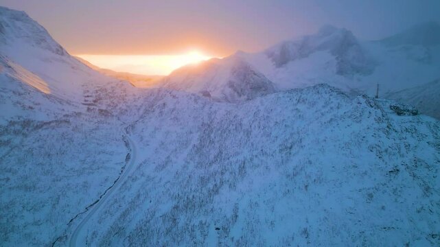 Sonnenaufgang in schneebedeckter Berglandschaft im Winter, klare Sicht auf die Sonne trotz Schneesturm in der Ferne, warmes Licht strahlt durch dunstige Atmosph&auml;re &uuml;ber den wei&szlig;en Gipfeln