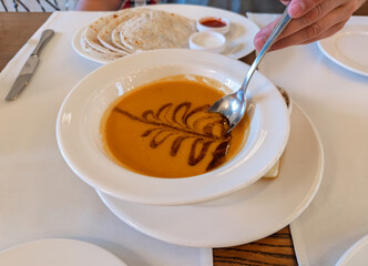 A person eating traditional Turkish red lentil soup, Mercimek Corbasi, garnished with spiced butter.First course of Turkish meal: A bowl of hot, creamy lentil soup being served at a restaurant table