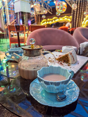 A glass teapot, a fancy blue cup of tea, and cake on a table in a restaurant with cozy, illuminated interior. Hot milk masala tea served in a beautiful glass teapot and light blue ceramic cup in cafe