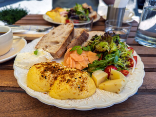 Gourmet breakfast platter with benedict eggs, salmon, fresh salad, cheese, and sourdough bread, served on a cafe terrace.A healthy and delicious brunch served al fresco on a wooden table with coffee