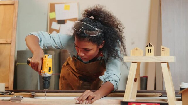 Carpenter woman using a power drill to secure a piece of wood in a workshop, focusing on precision in furniture making. Surrounded by tools and wood pieces, highlighting skill and craftsmanship
