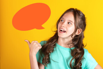 Portrait of cute young caucasian european blonde girl with opened mouth pointing thumb finger to the side side with empty blank orange speech bubble balloon dialog on yellow background. Studio shot