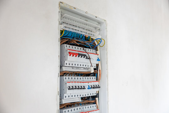 An open electrical panel embedded in a white wall, showing circuit breakers, blue, brown, and yellow green wires in an organized but unfinished state.