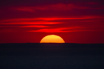 Red sunset over the sea horizon with the sun half-hidden behind a dark silhouette of distant hills.