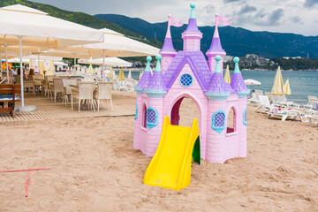 Pink plastic fairy-tale castle on beach sand close-up. Seaside playground with loungers, umbrellas, and mountains in background. Copy space