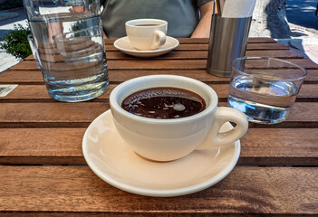 Morning Coffee on a Sunny Outdoor Cafe Terrace.Close-up of Americano Coffee and Water on a Wooden Table.Leisurely Afternoon at a Cafe: Two Cups of Coffee in the Sunlight
