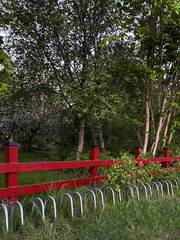 A vivid red fence with a silver bike rack placed in a green park surrounded by trees. Captures...