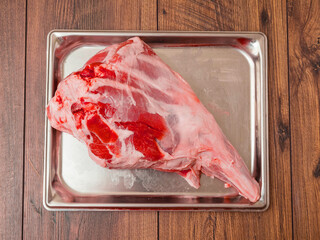 Fresh raw leg of lamb is on a silver stainless tray which is on a wooden table. Premium high...