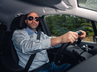 A man wearing sunglasses and a blue shirt is driving a car. He is wearing safety seat belt and has...