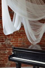 Old black grand piano on white floor in a loft interior with red brick wall and hanging white fabric. Concept: classical music, creativity, vintage style, artistic atmosphere.