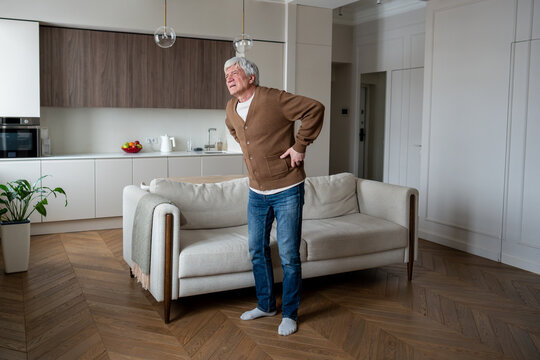 Senior man holds injured lower back, standing near couch in pain suffering. Lonely elderly male suffer from degenerative disc disease, spinal stenosis, chronic sciatica, mobility impairment. 