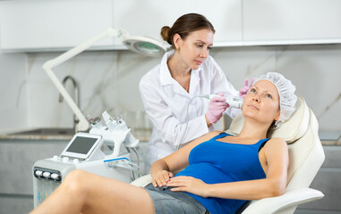 Obraz premium Young beautician with adult female client during hardware facial procedure in clinic. Concept of professional cosmetology and rejuvenation