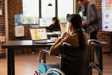 Businesswoman in wheelchair with clipboard and telephone in hand, seated at desk with laptop...